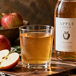 A glass of clear apple juice beside a bottle with a plain label and fresh red apples on a wooden table