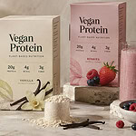 Three plain boxes labeled Vegan Protein in chocolate, vanilla and berry flavors on a stone counter, with a bowl of cocoa-brown powder, a scoop of pale powder, vanilla pods, berries and a pink smoothie