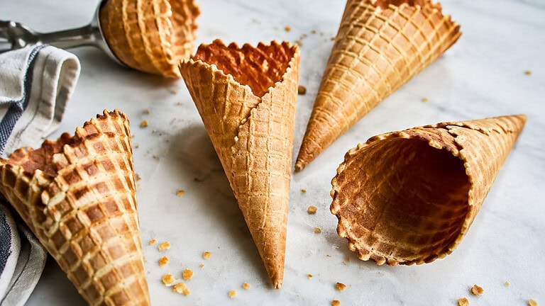Five empty golden waffle cones lying on white marble among a few crumbs, beside a metal ice cream scoop and a striped linen towel