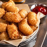 Golden crispy tater tots heaped on parchment in a metal basket on a wooden board, with a small cup of ketchup beside them