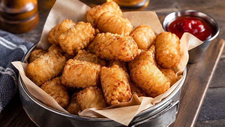 Golden crispy tater tots heaped on parchment in a metal basket on a wooden board, with a small cup of ketchup beside them