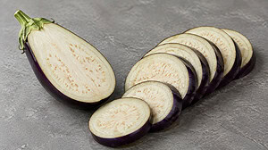 Half a raw eggplant cut lengthwise beside a fanned row of round eggplant slices on grey stone