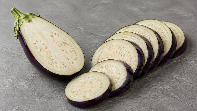 Half a raw eggplant cut lengthwise beside a fanned row of round eggplant slices on grey stone