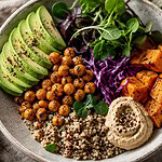 A grain bowl of sliced avocado, roasted chickpeas, quinoa, shredded red cabbage, roasted sweet potato cubes and a swirl of creamy dip, with a fork and a glass of water with lime beside it