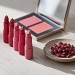 Five red and pink lipsticks and an open eyeshadow palette on a pale oak tabletop, next to a small stoneware dish of dark red dried cochineal insects in soft morning light.