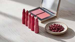 Five red and pink lipsticks and an open eyeshadow palette on a pale oak tabletop, next to a small stoneware dish of dark red dried cochineal insects in soft morning light.