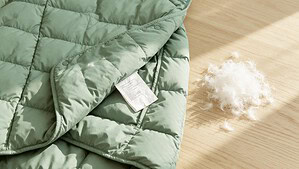 A sage green puffer jacket lies flat on a pale oak table with its inside care label folded out, beside a small loose pile of white goose down feathers.