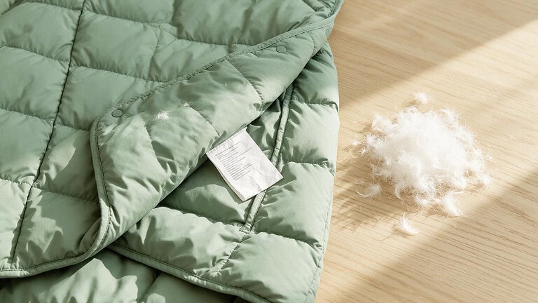 A sage green puffer jacket lies flat on a pale oak table with its inside care label folded out, beside a small loose pile of white goose down feathers.