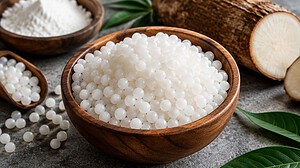 A wooden bowl of white tapioca pearls beside a bowl of tapioca flour and a halved cassava root on grey stone