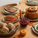 A Thanksgiving table set with a stuffed roasted pumpkin at the center, a sliced pastry-wrapped roast behind it, green beans with almonds, roasted carrots, sweet potato wedges, Brussels sprouts, cranberry sauce, a basket of rolls, small pumpkins and lit candles on a wooden table