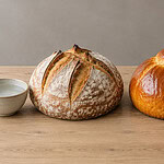 A flour-dusted round loaf with a bowl of water beside it, next to a glossy golden loaf with a jug of milk, an egg, a cracked egg yolk and a block of butter on a wooden table