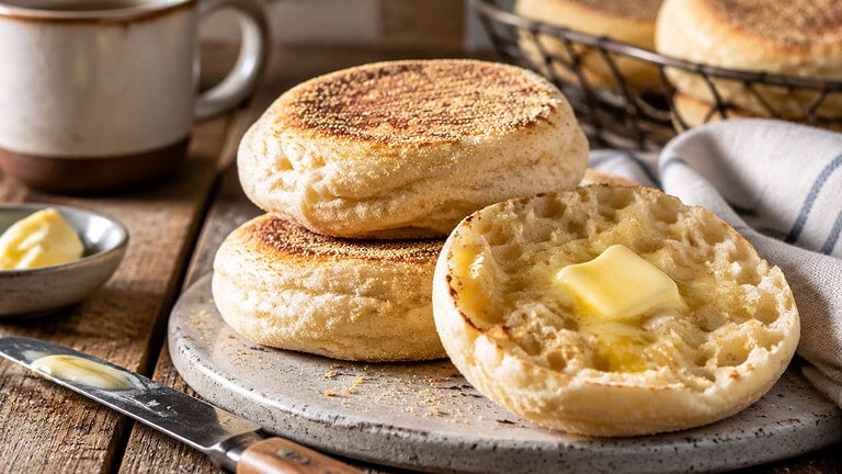 Toasted English muffins stacked on a stone board, one split open with a pat of melting spread, beside a small dish of spread, a knife, a mug and a linen cloth.