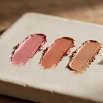Three swatches of cream makeup in rose, terracotta and warm bronze beside a small mound of golden-beige loose powder on a pale stoneware tile