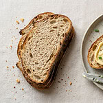 A slice of sourdough bread spread with pale yellow vegan butter and chopped chives on a speckled stoneware plate, a butter knife beside it and two more slices on a linen cloth